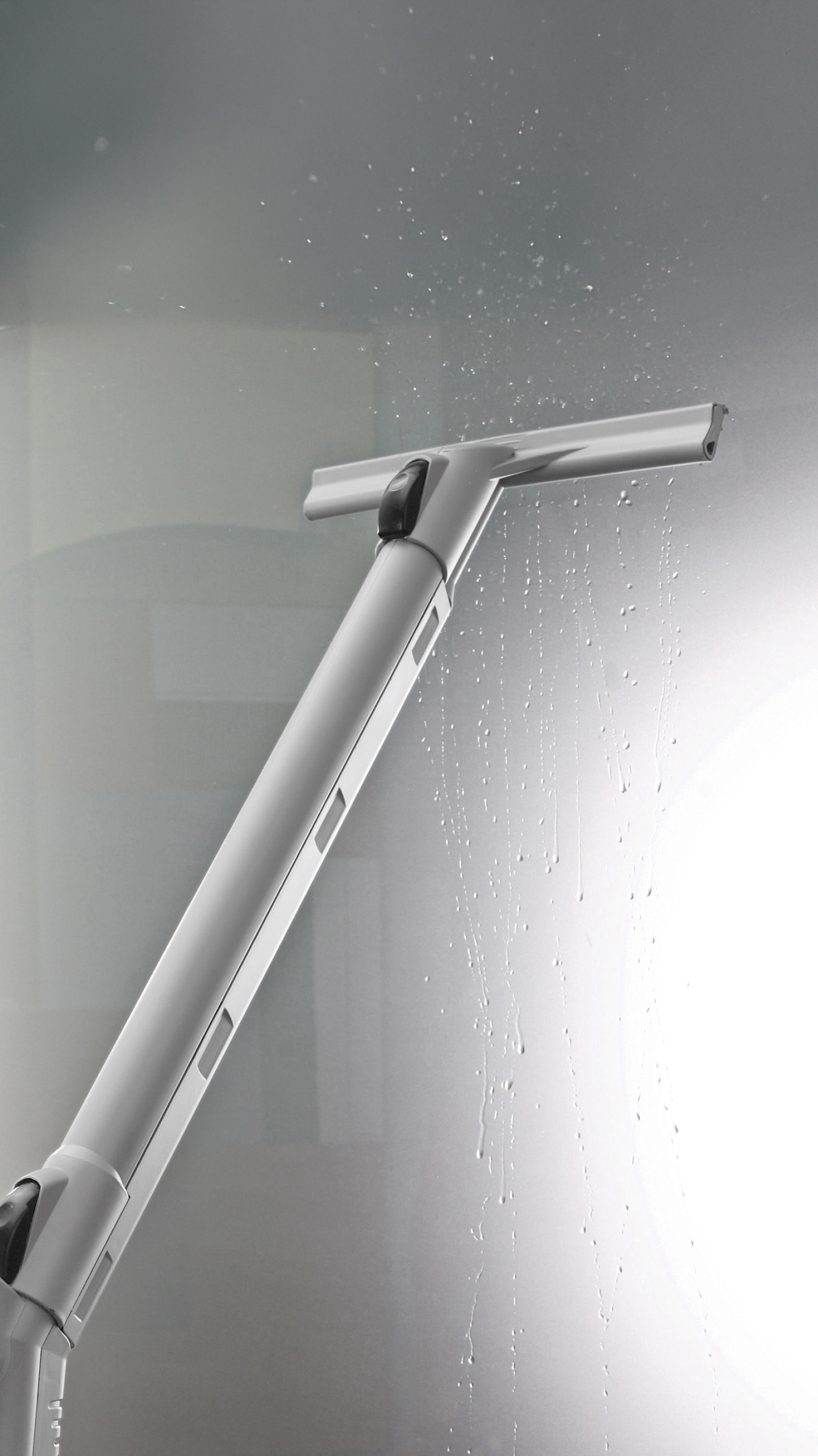 A close-up of a handheld squeegee cleaning a wet glass surface, with droplets of water visible. The squeegee features a long handle and a wide cleaning blade.